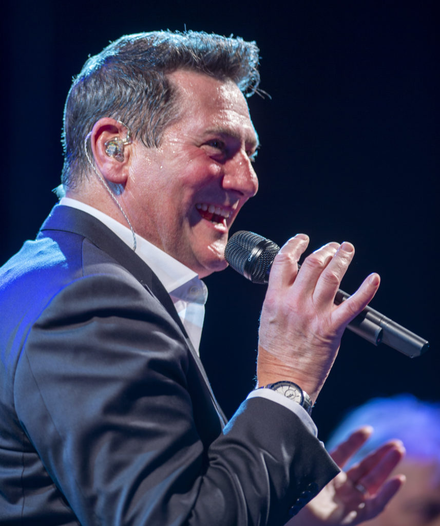 Tony Hadley sorride durante il concerto, microfono nella mano destra e auricolari; abbigliato con camicia bianca e vestito grigio