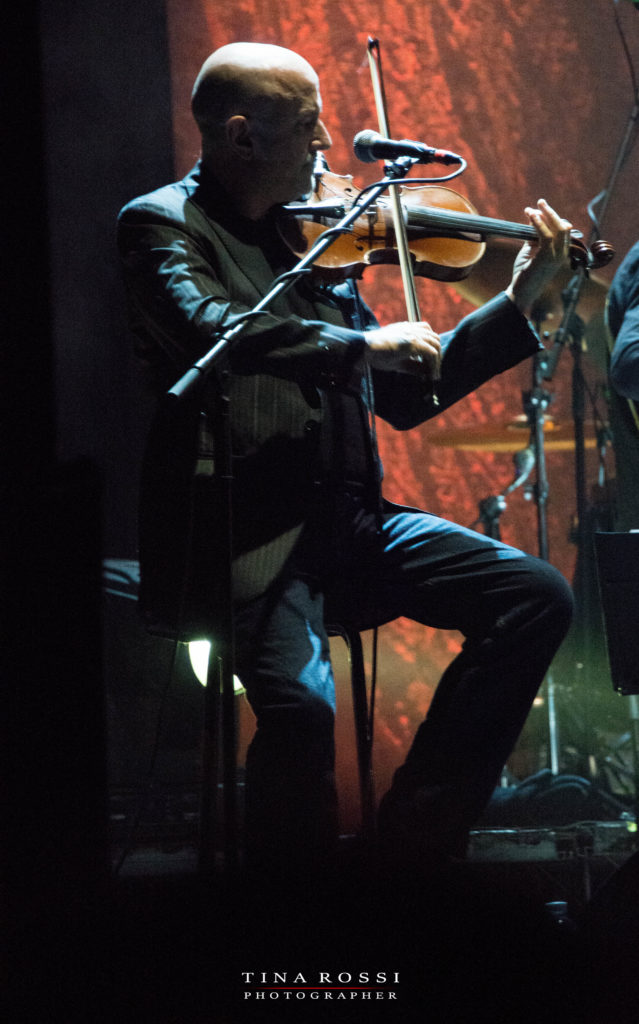 primo piano di lucio fabbri, seduto su uno sgabello, vestito di grigio, intento a suonare il violino