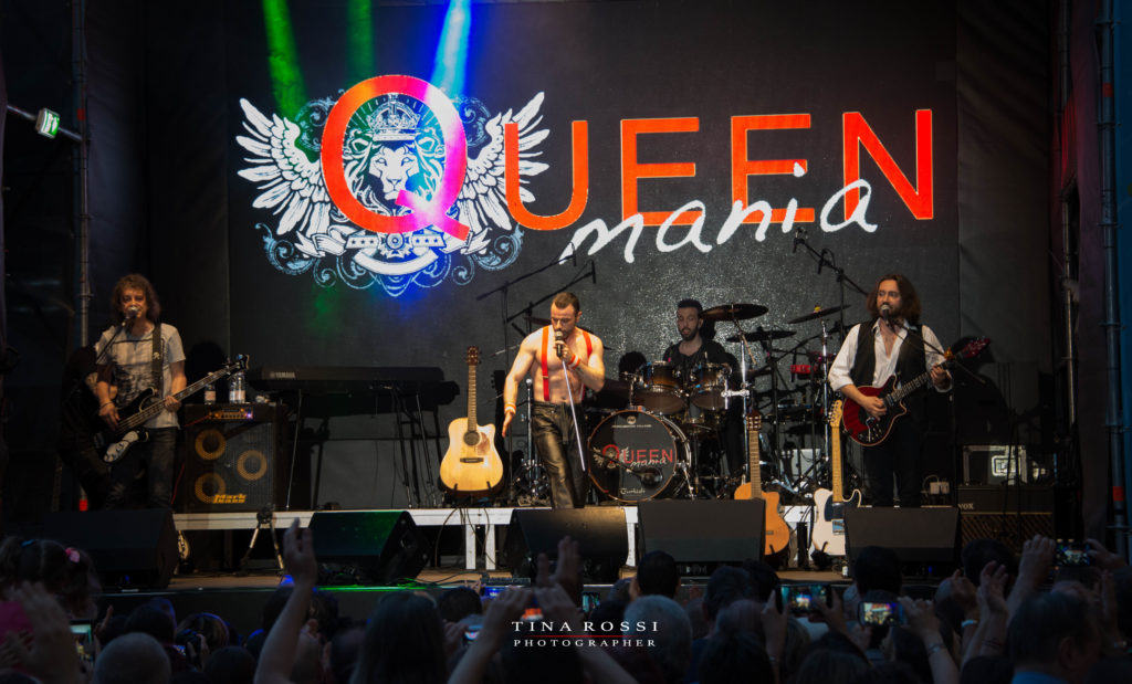 Vista del palco con la band che suona. La scritta "queenmania"grande sullo sfondo colorata di rosso e la gente sotto il palco che batte le mani