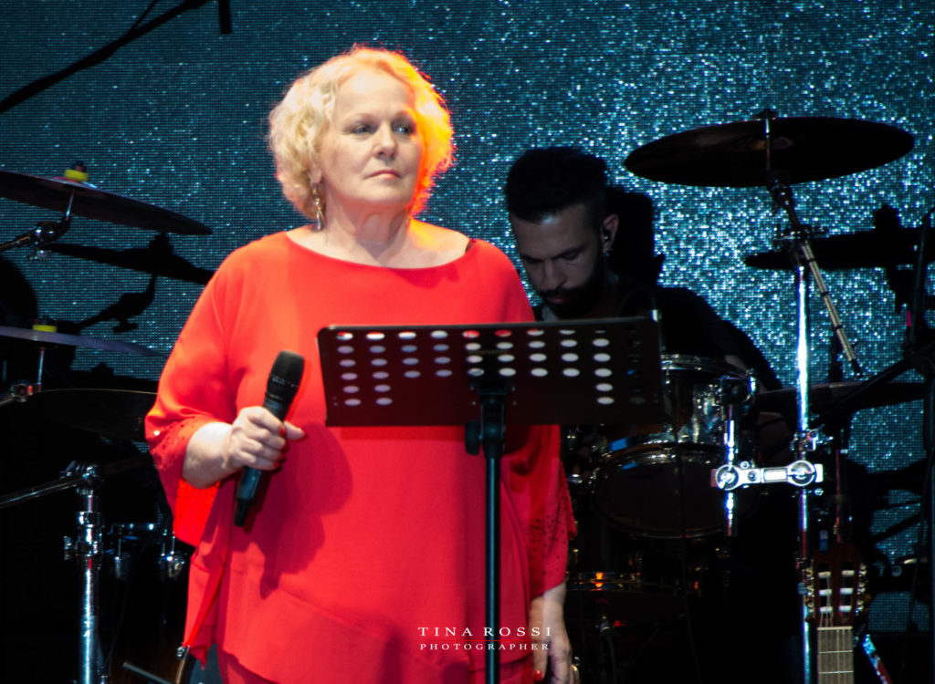 Katia Ricciarelli vestita con una casacca rossa a maniche lunghe, con il microfono in mano guarda assorta il pubblico Dietro di lei il batterista e la batteria
