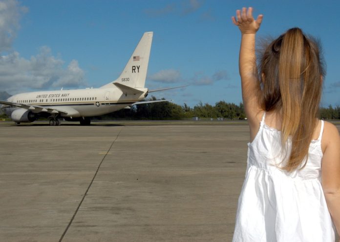 bambina vestita di bianco, girata di schiena, con i capelli biondi raccolti in coda di cavallo, sulla pista di un aereoporto che saluta un aereo