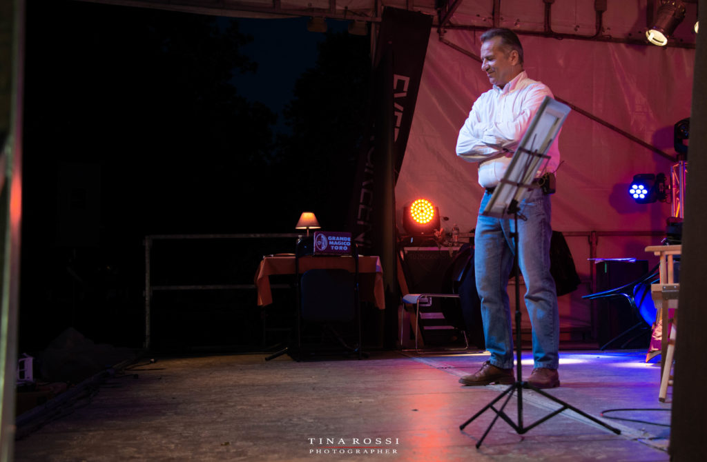 Vincenzo Santagata vestito con jeans e camicia bianca con le braccia conserte sul palco con sullo sfondo una scrivania con una lampada accesa e il cuscino con scritta grande magico Toro