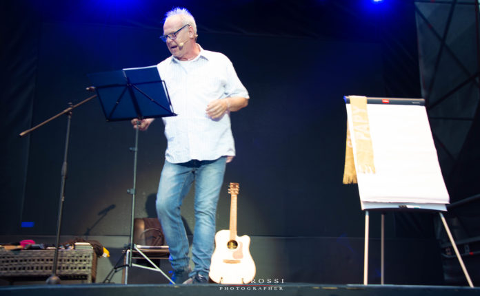 Pino Campagna sul palco con gli occhiale legge su un leggio, affianco a lui una chitarra e una lavagna