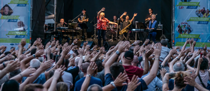 Nino Frassica vestito di rosso con la Los Plaggers Band sul palco e un folto pubblico che agita le mani