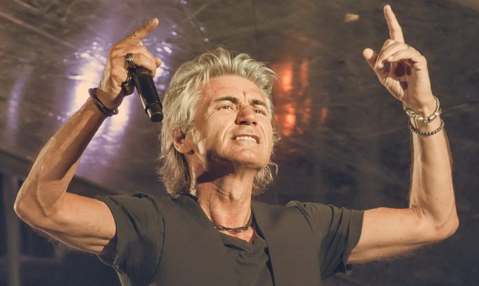 Ligabue vestito con una t shirt marrone, capelli lunghi con le mani verso il cielo