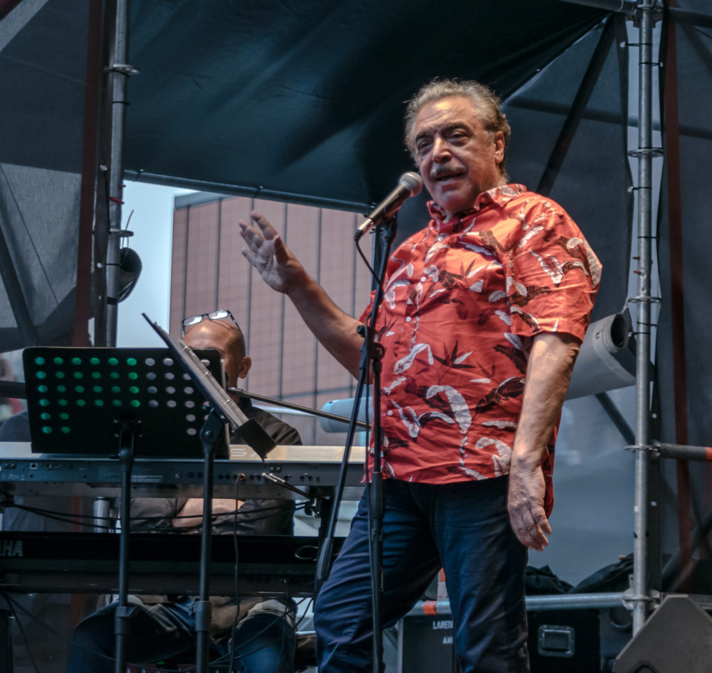 Nino Frassica con camicia rossa a fiori, parla davanti al microfono, sorridendo al pubblico, di fianco a lui un leggio