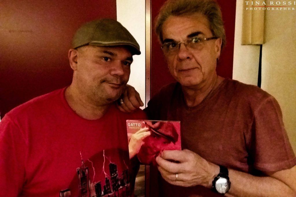 Primo piano di Gatto Panceri e Lele Boccardo, entrambi indossano una t-shirt rossa e Lele tiene in mano la copertina del dc Pelle d'oca e lividi