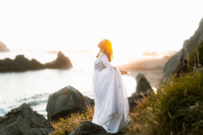 respiro nel passato - donna vestita di bianco sugli scogli con il mare sullo sfondo , che guarda verso il cielo illuminata dai raggi del sole