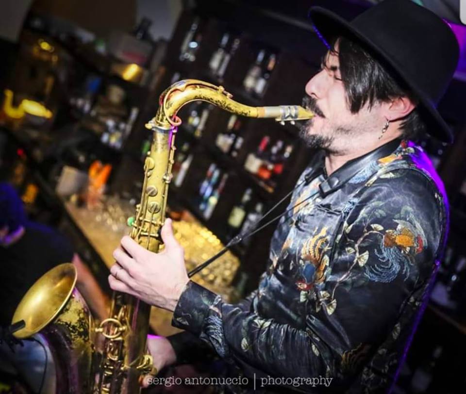 daniele vitale di profilo con una bombetta in testa e una camicia nera a fiori suona il sax