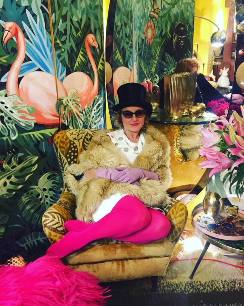 Monica Bruno titolare di Marco Polo, seduta su un divano indossa fuseaux fucsia , una pelliccia, un cppello e occhiali neri. Sullo fondo pannelli con fenicotteri rosa