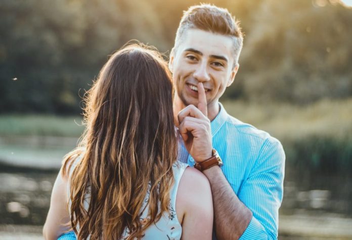 coppia abbracciata, lei di spalle e lui sorridente si mette un dito sulle labbra in segno di silenzio