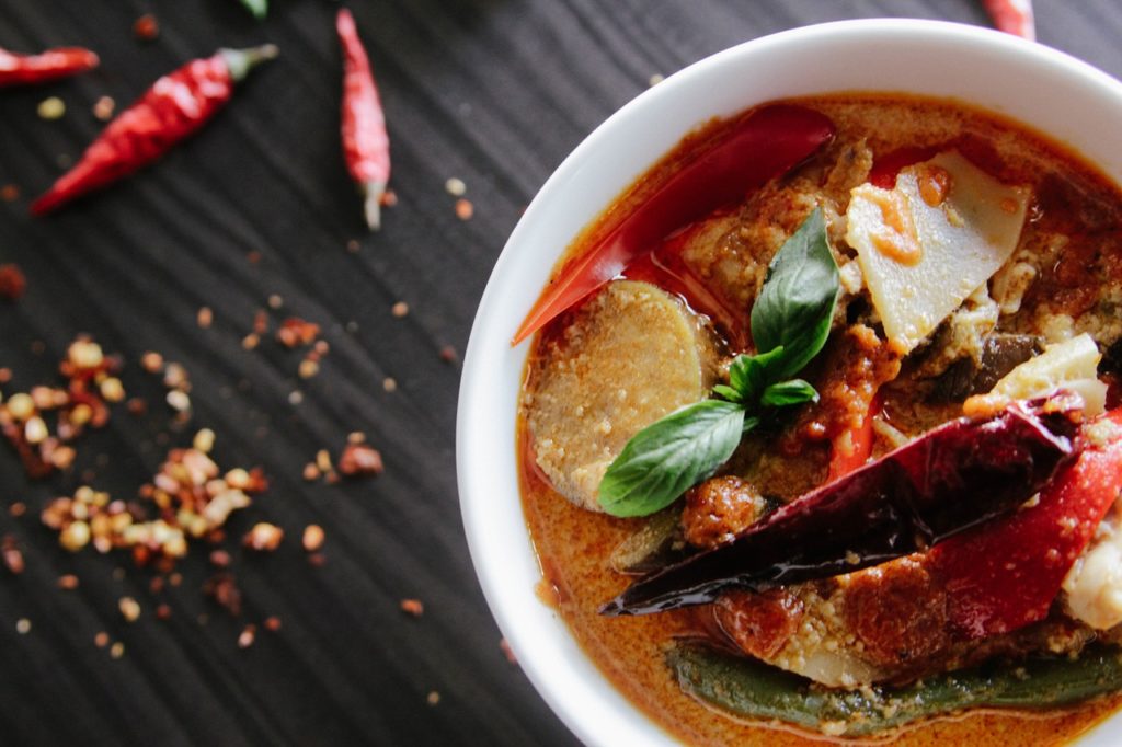 una zuppa con verdure e peperoni di carmagnola in una ciottola bianca appoggiata su un tavolo di legno dove ci sono peperoncini e semi 