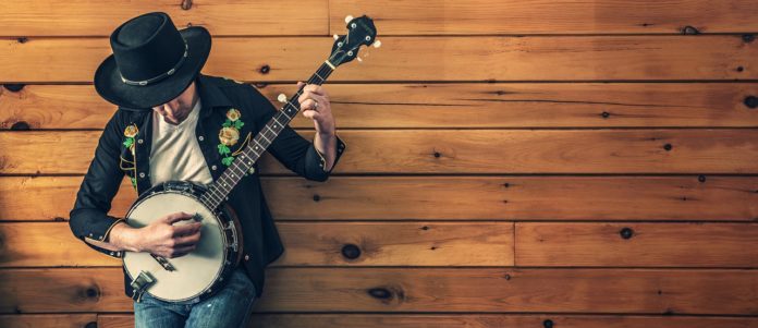MR. SIL al Gv pane e caffè, in primo piano un suonatore di banjo con un cappello nero da cawboy, sullo sfonto un muro di assi di legno