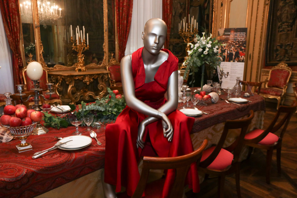 Un manichino con abito da sera rosso è seduto su un tavolo antico apparecchiato, e con i piedi appoggiati su una sedia e le braccia appoggiate sulle ginocchia. E' parte della collezione di Iconica 2019