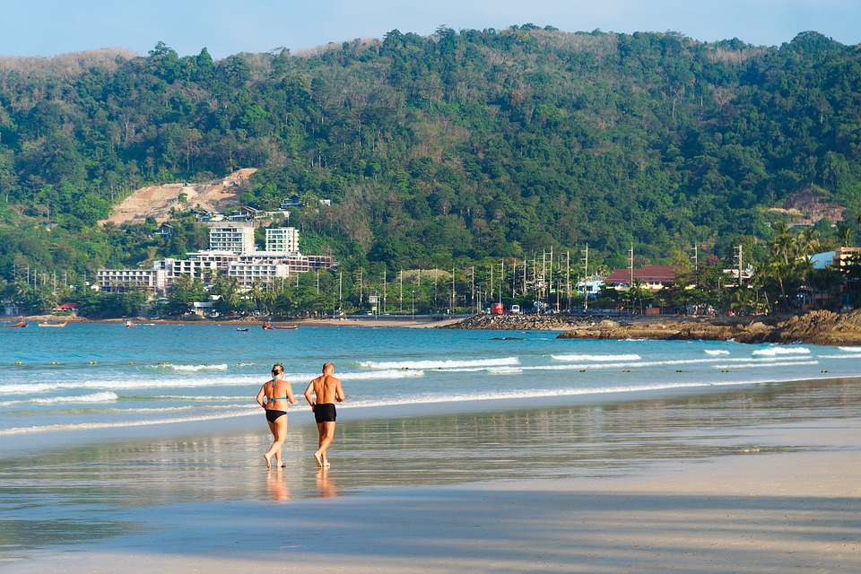 una coppia in costume da bagno, di schiena fa footing su una spiaggia, sul bordo del mare. Sullo sfondo un paese con una montagna