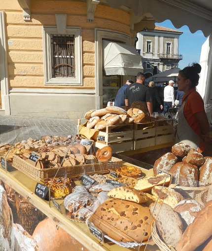 Una bancarella con del pane in vendita e una signora dietro le ceste con grembiule scuro e maglia rossa