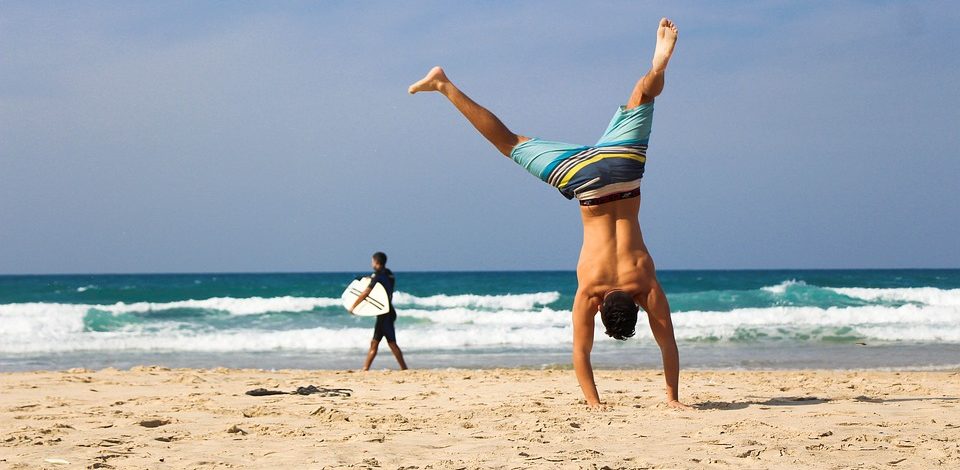 allenarsi in vacanza : Su una spiaggia un uomo in primo piano fa la verticale sulla spiaggia e uno sullo sfondo con una tavola da surf sotto il braccio