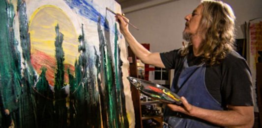 il falsario d' arte visto lateralmente, con una maglietta nera mentre dipinge con la tavolozza in mano un quadto molto grande con verdi e azzurri