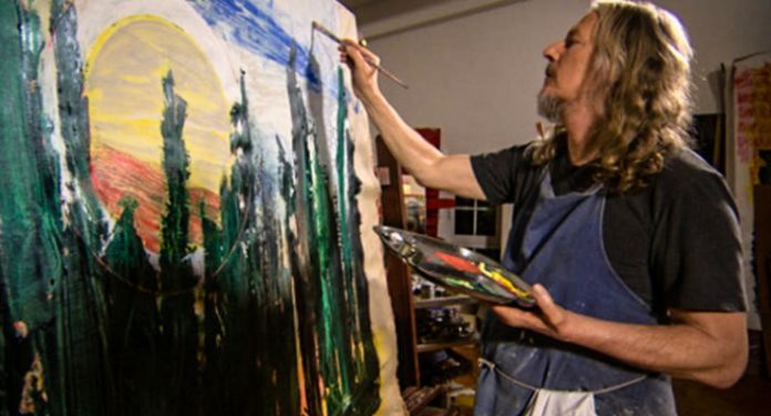 il falsario d' arte visto lateralmente, con una maglietta nera mentre dipinge con la tavolozza in mano un quadto molto grande con verdi e azzurri