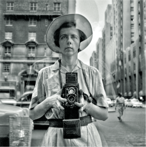 Ritratto in bianco e nero di Vivian Maier, con macchina foografica in mano. Indossa un cappello,e sullos fondo la città