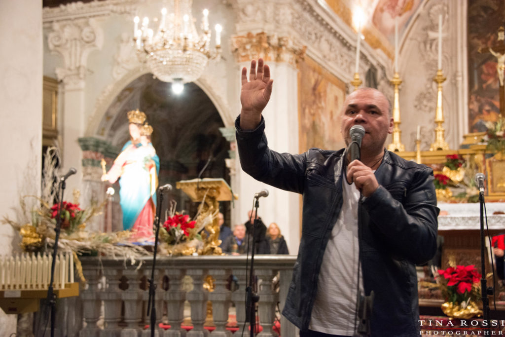 Gatto Panceri con il braccio destro alzato e il microfono nella mano sinistra sta cantando in una chiesa e sullo sfondo la statua della Madonna