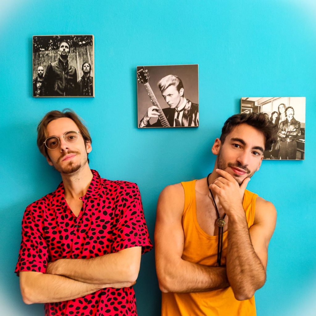 Le Tendenze: Daniele e Francesco Saibene, i cantanti de la vita di un clown.  Daniele è vestito con una camicia leopardata rossa e nera e Francesco con una canottiera gialla. Dietro di loro su una parete azzurra il quadro di David Bowie