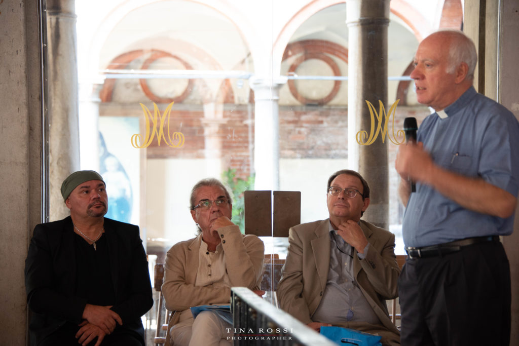 Padre Lucchina, in piedi, parla al pubblico, seduti vicino a lui Gatto Panceri, lele Boccardo e Enzo Toniutto, mentre il parroco spiega la storia di una piccola Lourdes
