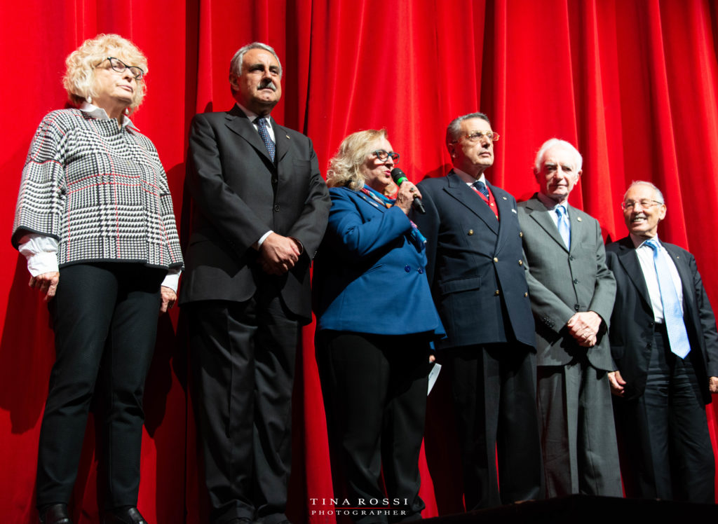 La Governatrice Matrotisi con tre membri Rotariani infila sul palco di un teatro con dietro il sipario rosso e loro sono vestiti da gran galà