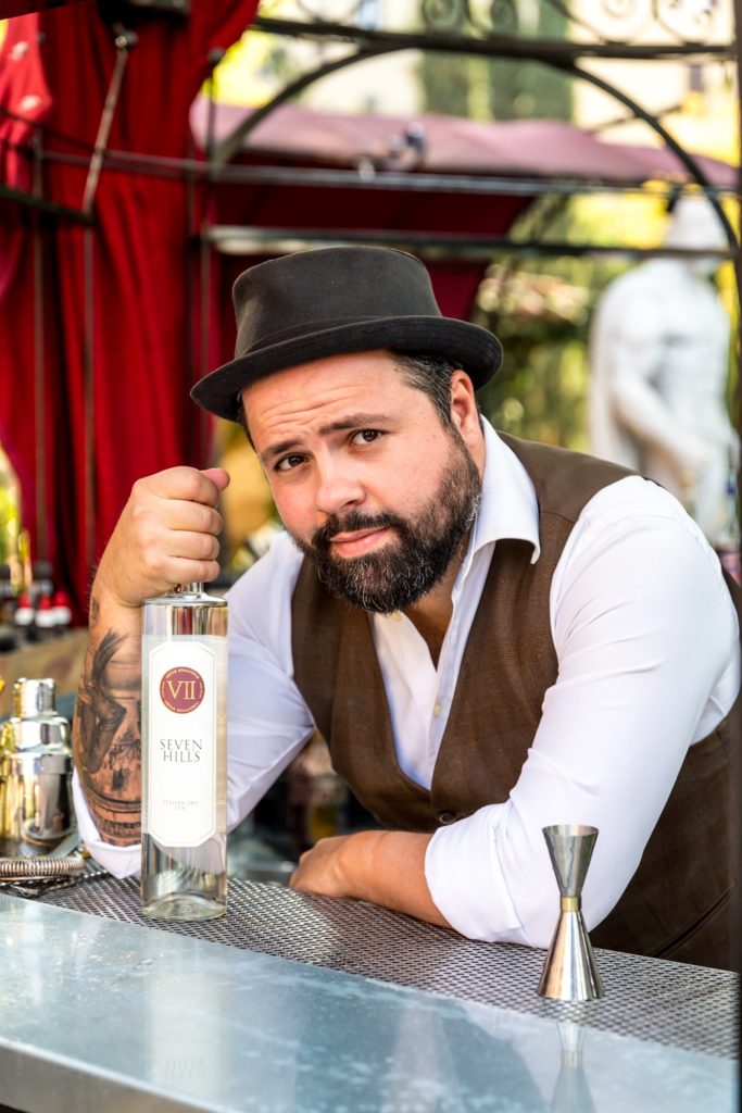 Federico Leone con un cappello , camicia bianca e gillet marrone appoggiato su un tavolo tiene in mano una bottiglia di gin con la quale preparerà la drink list