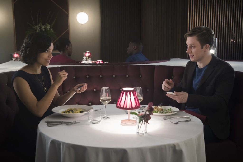 Black mirror: Una coppia seduta ad un tavolo di ristorante, rotondo, con ciascuno davanti un piatto di pasta un bicchiere di vino bianco e una lampada rossa a centro tavola, stanno sorridendosi e chiacchierano