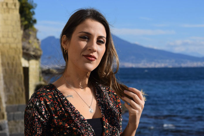 Cristina Cafiero: primo piano della cantante, che indossa un vestito a fiori. Sullo sfondo il mare e il vesuvio