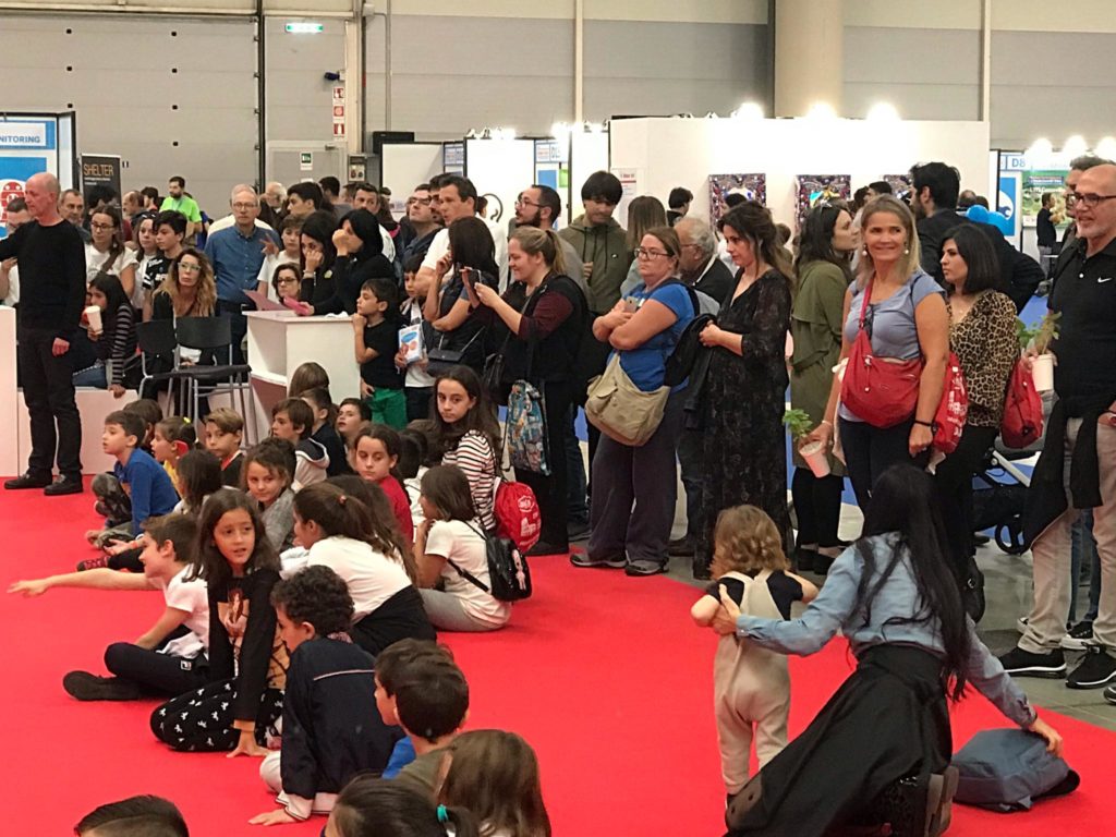 pubblico presente alla maker faire. bambibi seduti su tappeto rosso e molti adulti in piedi che seguono una dimostrazione