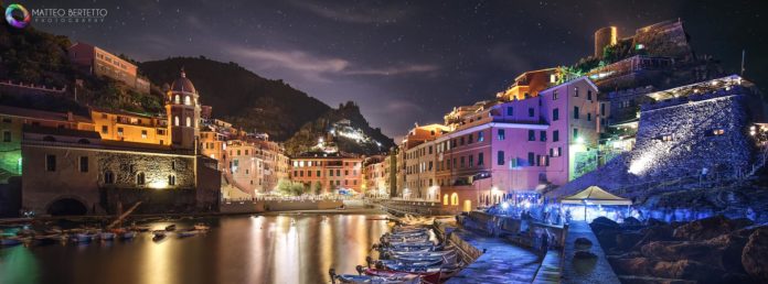 una fotografia moderna con un panorama notturno di un paese illuminato che si specchia nel mare, in primopiano il porto con le barche attraccate, realizzata con una tecnica particolare per far risaltare le luci