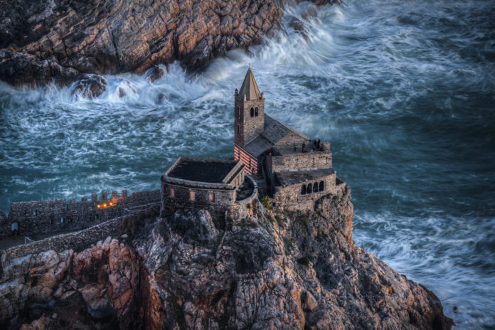 Una veduta dall'alto di San Pietro in Porto Venere, realizzata con la tecnica di fotografia moderna di Matteo Bertetto, una rocca con in cima una castello e il mare in burrasca intorno