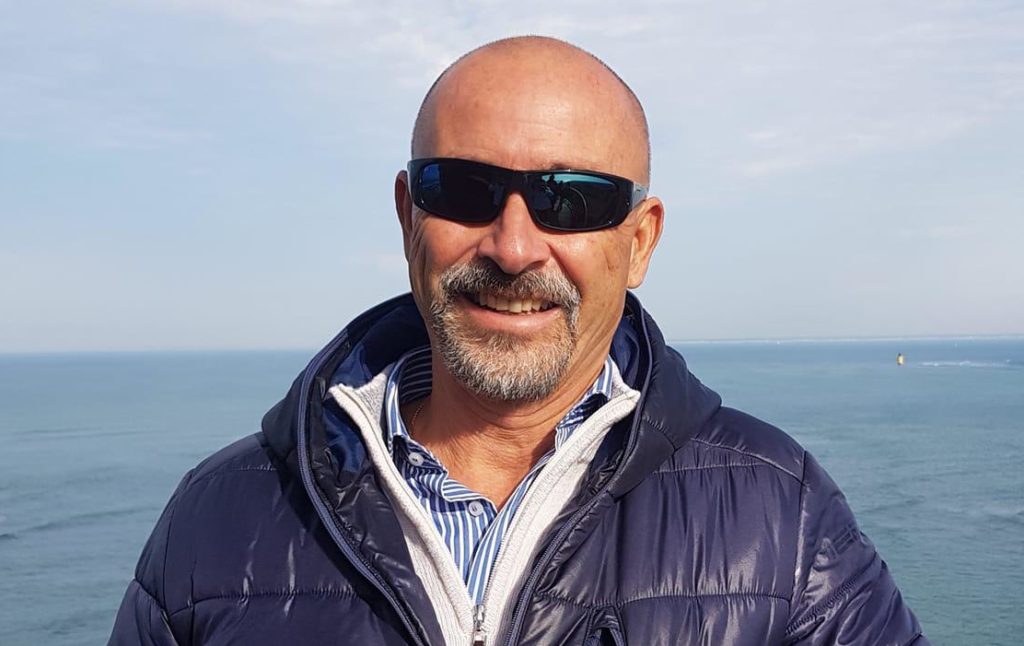 Americo Pilati, presidente di federalberghi liguria, calvo, con occhiali da sole neri, sorridente, con pullover bianco e piumino blu, sullo sfondo il mare