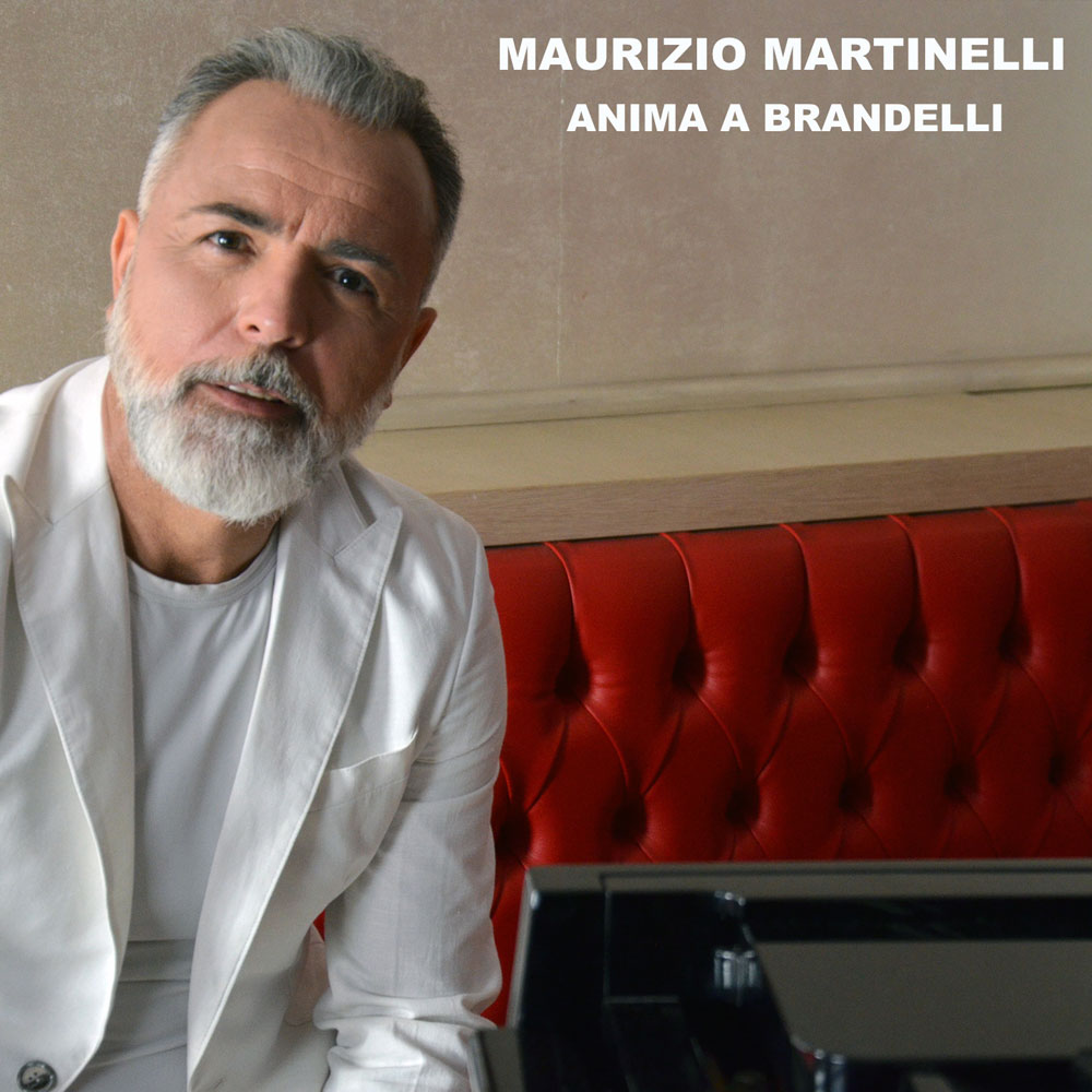 i dettagli di un giorno " di Maurizio Martinelli rappresentato seduto su un divano di pelle rossa. il cantautore indossa una giacca e una maglietta bianche