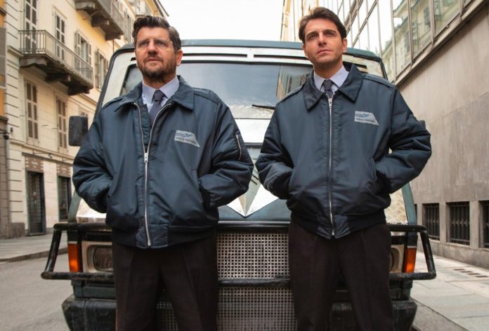 Gli uomini d'oro: primo piano dei protagonisti Fabio De Luigi e Giampaolo Morelli, in divisa da dipendenti delle poste, appoggiati ad un furgone postale
