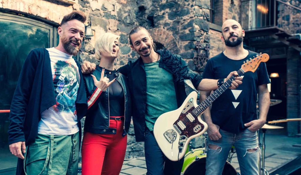 gli sugarfree in una foto di gruppo con Serena De Bari nella copertina del nuovo singolo Frutta. La foto è scattata in un cortile con dietro un muro di mattoni. Mattia tiene in mano una chitarra