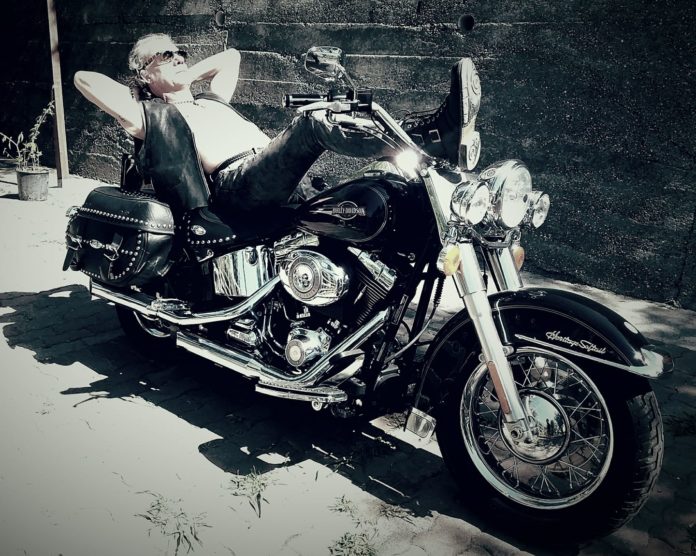 Harleysta: un uomo, che indossa gilet di pelle, jeans neri e stivali neri, sdraiato sopra una Harley Davidson nera
