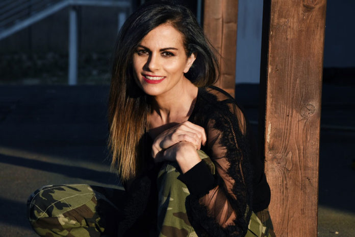 Antonella Bucci seduta con le braccia appoggiate su un ginocchio, sorride all'obiettivo. Indossa un'elegante maglietta nera e un paio di pantaloni militari.