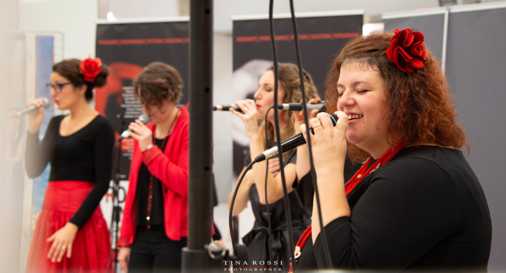 Tre componenti del gruppo vocal boutique una bionda in primo piano e le altre due more vestite di nero cantano a cappella