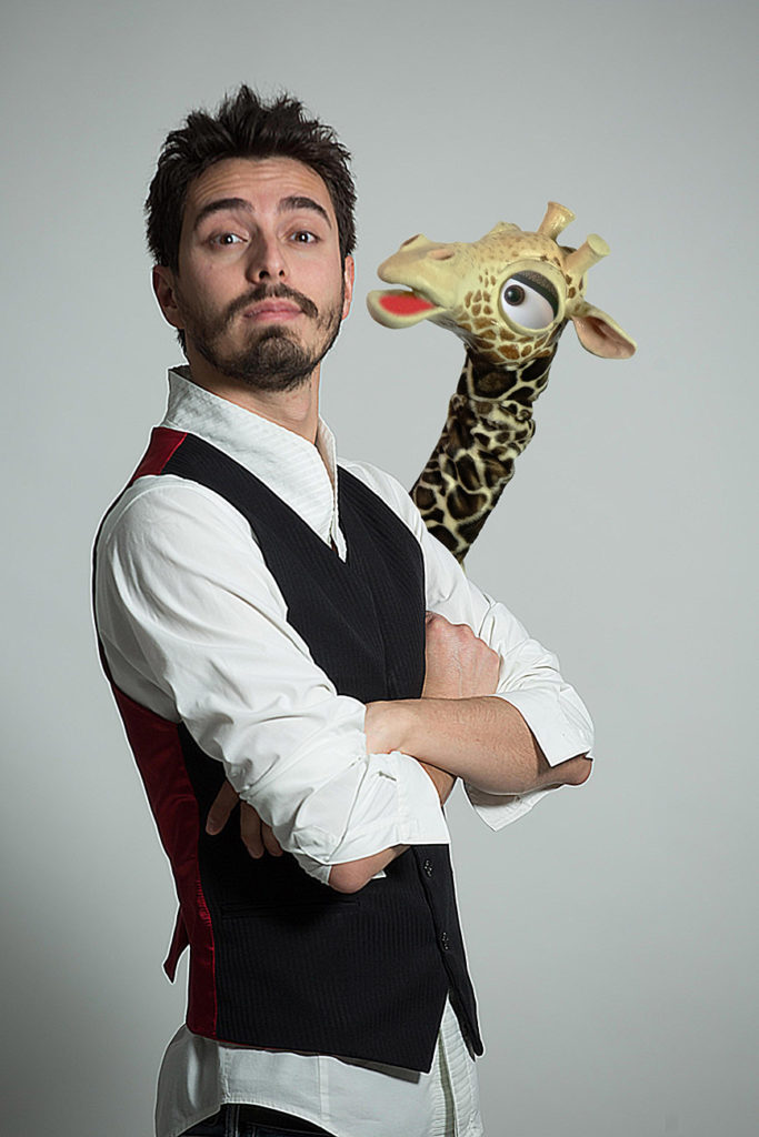 Rafael Voltan in primo piano a braccia conserte e sguardo serio, in camicia bianca e gilet nero, alle sue spalle spunta il collo della giraffa Gi