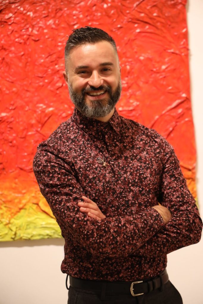 Massimiliano Ferragina, artista della mostra, sorride mostrando i denti, in primo piano, veste una camicia bordeaux a fiorellini e pantaloni neri, foto scattata da Marco Tanfi