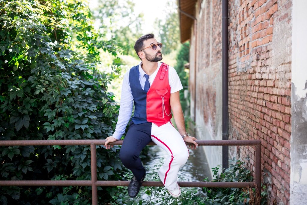 abbiamo preso l'influencer, di Riccardo Inge: primo piano del cantautore milanese, seduto su una ringhiera, che indossa pantaloni blu e bianchi, e una camicia bianca sotto un gilet blu e rosso