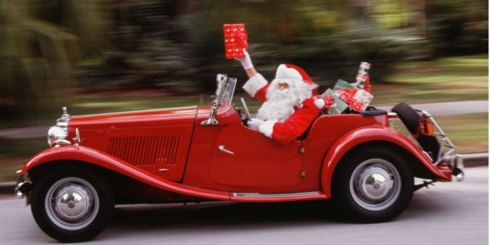 Babbo Natale che guida una macchina d'epoca rossa, con in mano un pacchetto regalo. Il cofano della macchina è piena di pacchetti regalo.