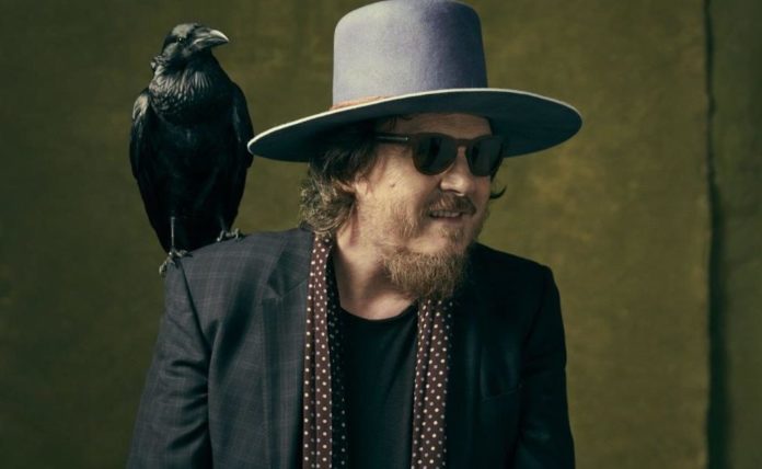 ZUCCHERO D.O.C. con un cappello a larga falda, vestito scuro, occhiali da sole scuri e un corvo sulla spalla