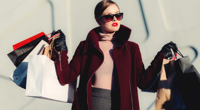 una donna con la sindrome da shopping, vestita elegante, con occhiali neri, maglioncino beige e le mani piene di pacchetti acquistati con il black friday