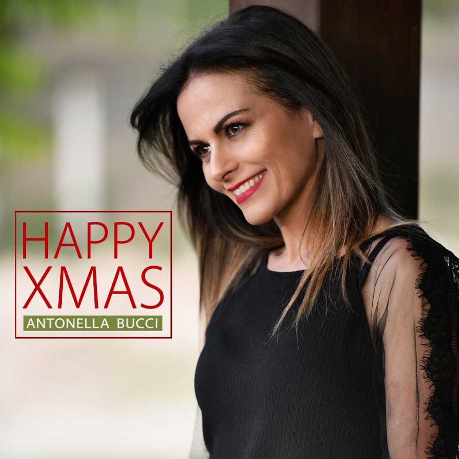 Antonella Bucci in primo piano, mezzo busto, con lo sguardo rivolto verso sinistra, dove c'è scritto Happy Xmas in rosso, . Indossa una maglietta elegante nera, e i capelli sono sciolti, che cadono sulle spalle