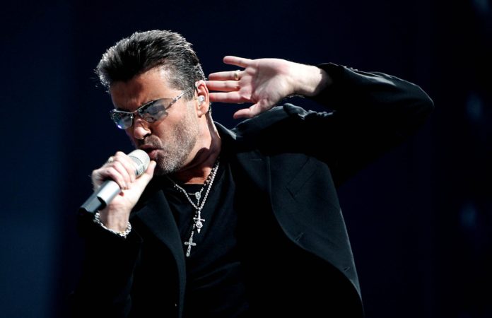 last xmas george michael canta last xmas vestito con maglione scuro, una croce al collo occhiali scuri e tienne la mano vicino all'orecchio mentre con l altra tiene un microfono