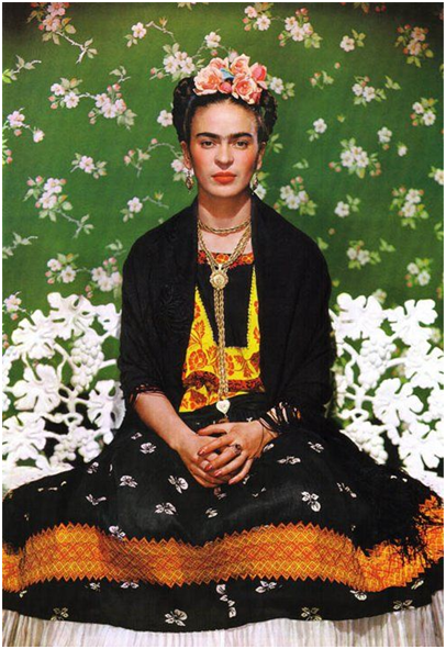Frida Kahlo in primo piano seduta con abito nero e giallo, capelli raccolti in una corona di rose rosa su sfondo verde con fiori bianchi, una foto presente nella mostra sulla sua vita